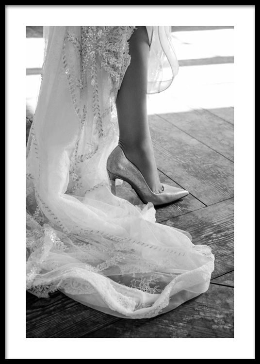  – Black and white photograph of the end of a white dress with embroidery and a high heeled shoe