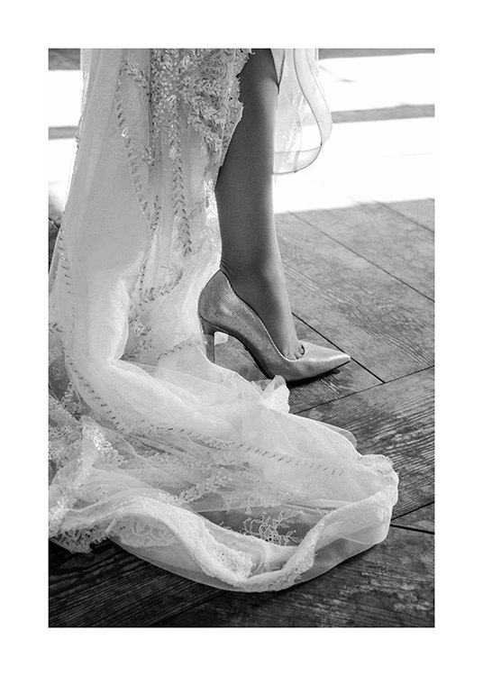  – Black and white photograph of the end of a white dress with embroidery and a high heeled shoe