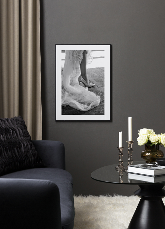  – Black and white photograph of the end of a white dress with embroidery and a high heeled shoe